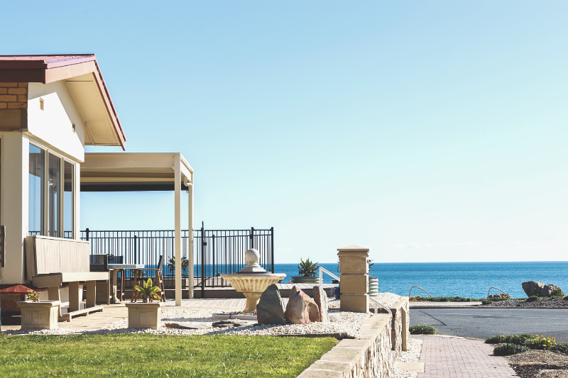 A modern house with large windows and a patio overlooks a calm blue ocean. The yard has a fountain, rocks, and green grass, with a clear sky above and a paved walkway leading toward the water.